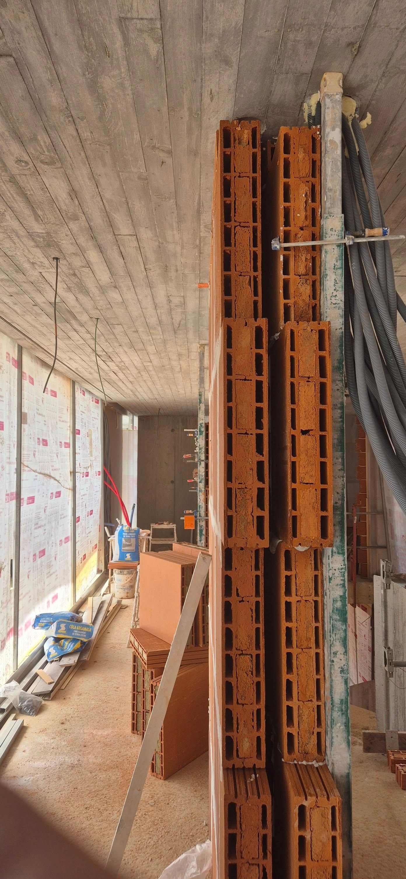 Couloir de chantier avec briques orange empilées, plafond en béton apparent, murs inachevés et câbles apparents.