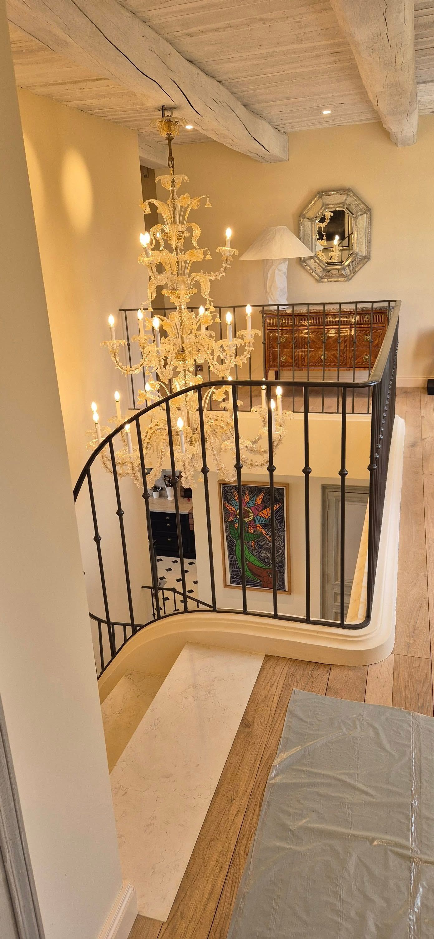 Palier d'escalier avec rampe noire, lustre et miroir orné dans un intérieur à l'éclairage chaleureux.