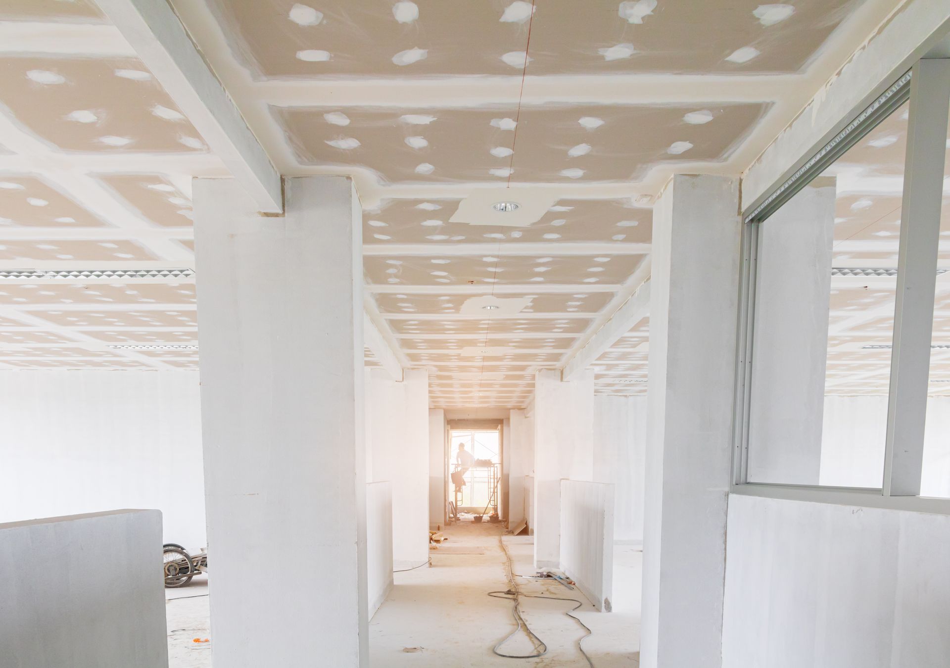 Couloir lumineux inachevé avec murs et panneaux de plafond blancs, fenêtres à droite et un câble au sol.