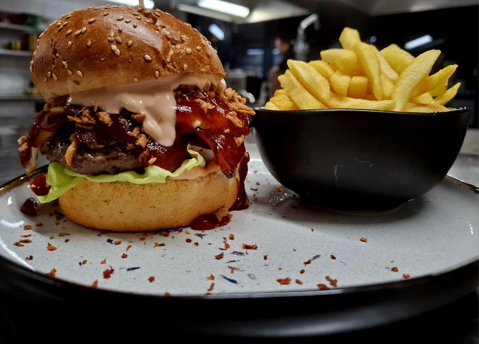 Un hamburger avec du bacon et des frites dans une assiette, accompagné d'un bol de frites noires.
