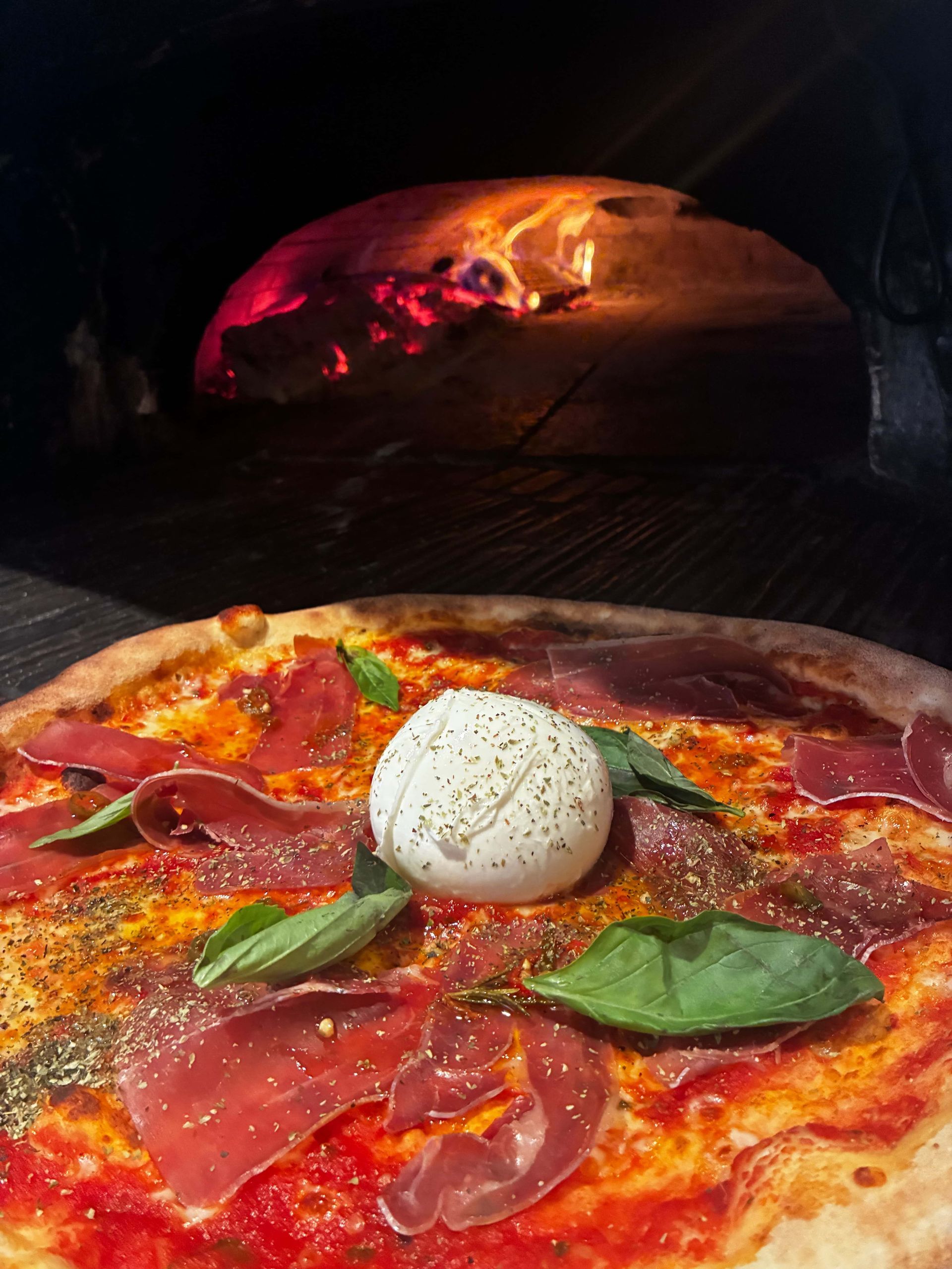 Pizza avec sauce tomate, basilic, pepperoni et une boule de burrata devant un four à bois