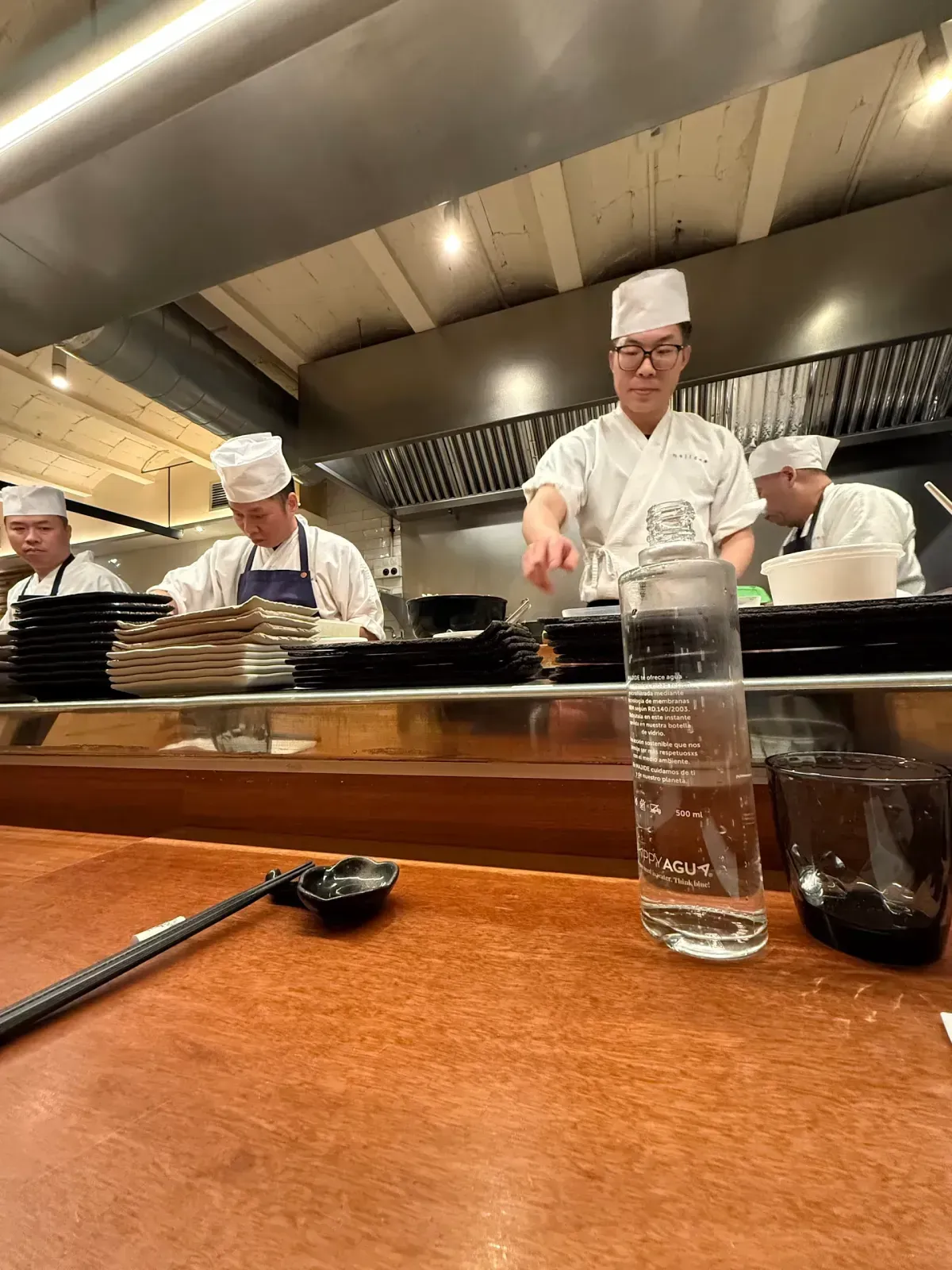 Chefs de sushi preparan comida tras un mostrador de madera. Un chef señala, mientras otros trabajan con platos en la cocina
