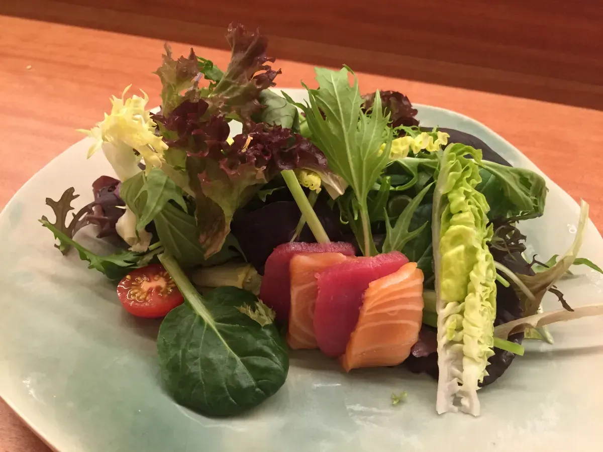 Ensalada de salmón crudo en rodajas y atún, con verduras mixtas y un tomate cherry, en un plato azul claro.