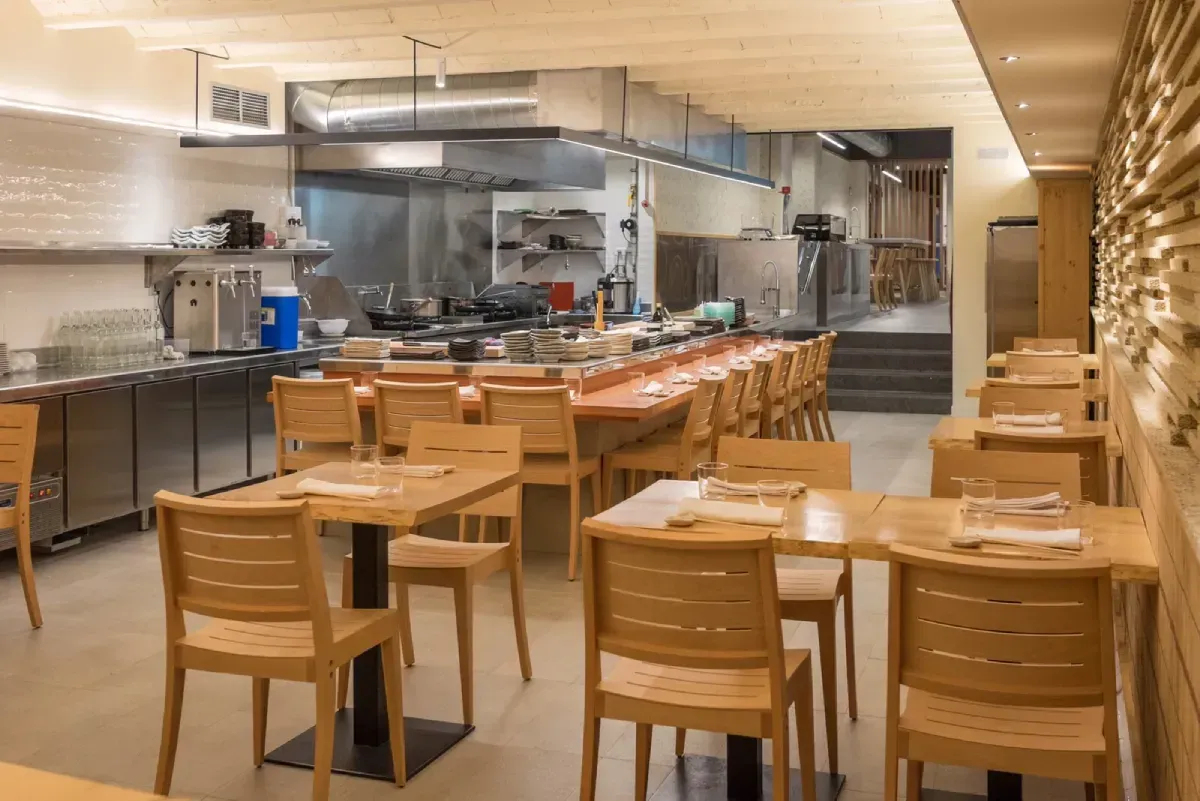 Interior de restaurante con mesas y sillas de madera, cocina abierta y paredes en tonos neutros.