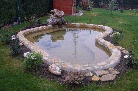Un estanque de jardín con forma de riñón, bordeado de piedra, ajardinado circundante y una pequeña roca en medio de un césped.