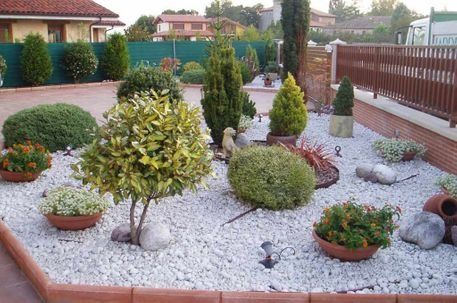 Un jardín de rocas con arbustos perennes variados, flores en macetas y grava decorativa blanca, bordeado por baldosas de terracota.