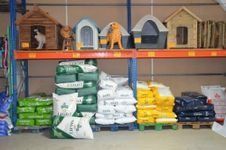 En una tienda, se ven estantes con varias casetas para mascotas en la parte superior y pilas de bolsas de comida para mascotas de diferentes colores en el suelo.