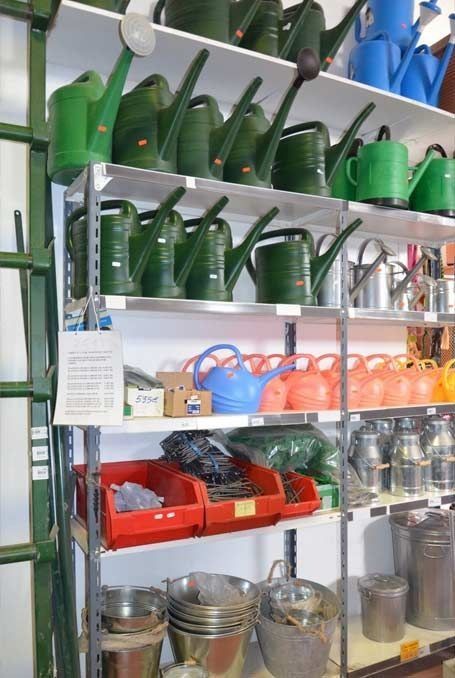 En los estantes de una tienda de jardinería se exhiben regaderas de plástico de color verde y naranja, cubos de metal y diversos accesorios de ferretería.