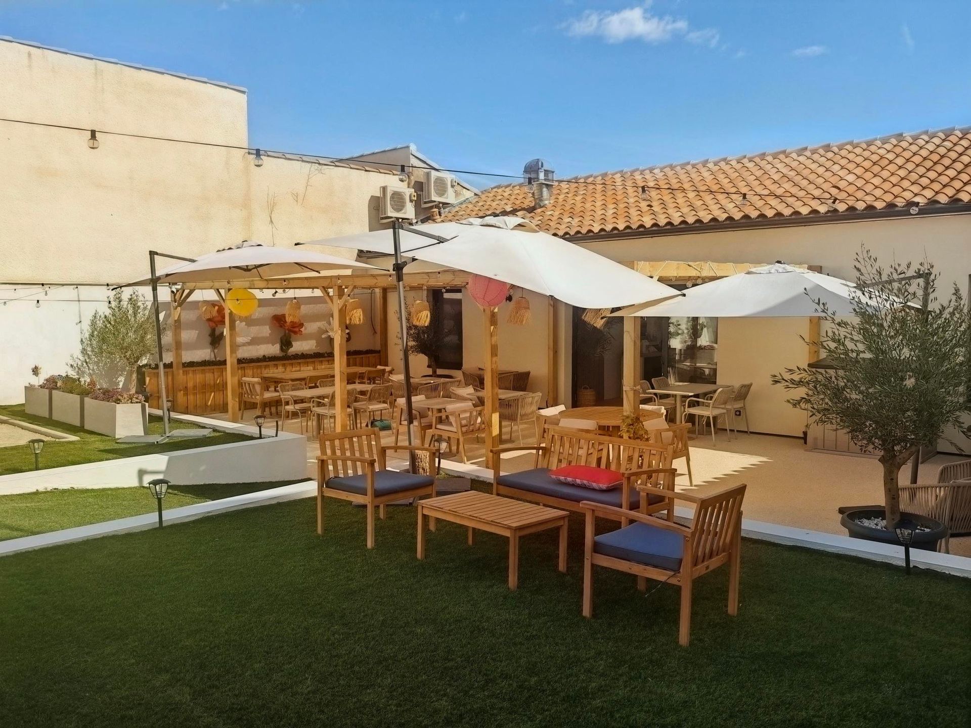 Terrasse extérieure avec tables en bois, parasols blancs et chaises rembourrées sur une pelouse verte près d'un bâtiment