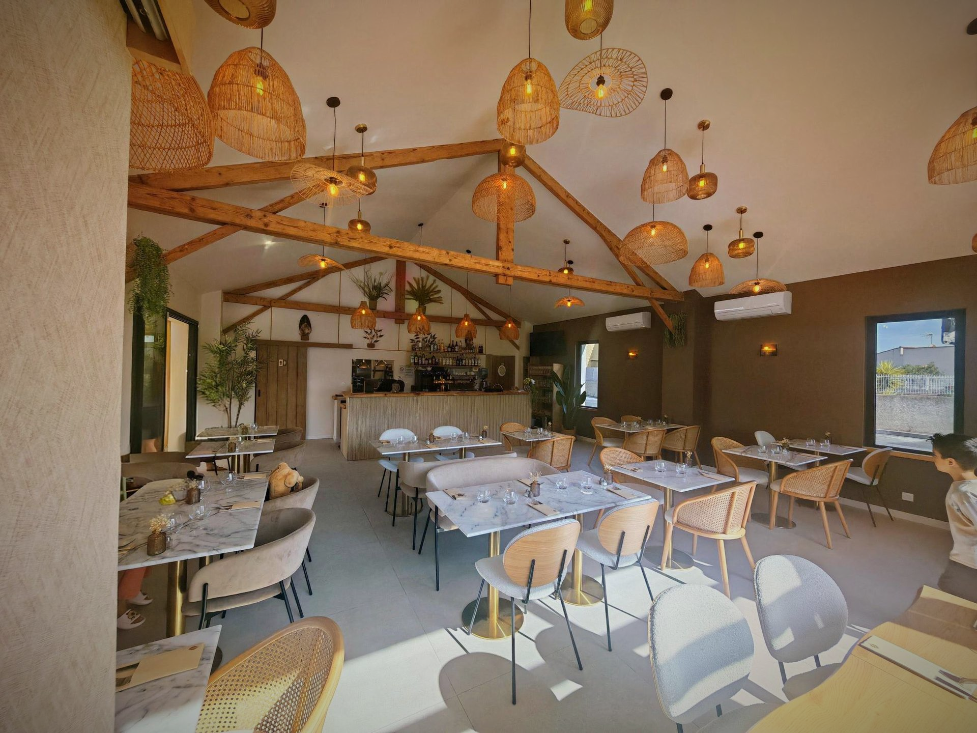 Salle à manger lumineuse et rustique avec tables blanches, chaises en bois et suspensions lumineuses en osier sous poutres apparentes.