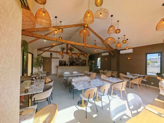 Salle à manger lumineuse du restaurant avec poutres apparentes, luminaires suspendus et tables dressées près de grandes fenêtres.