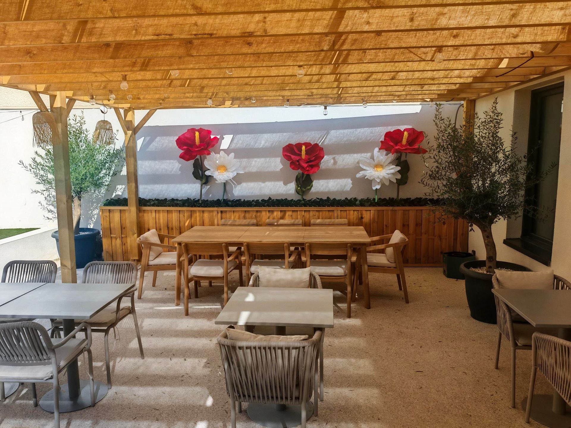 Espace repas extérieur couvert avec tables et chaises en bois, décorations florales rouges et blanches et plantes