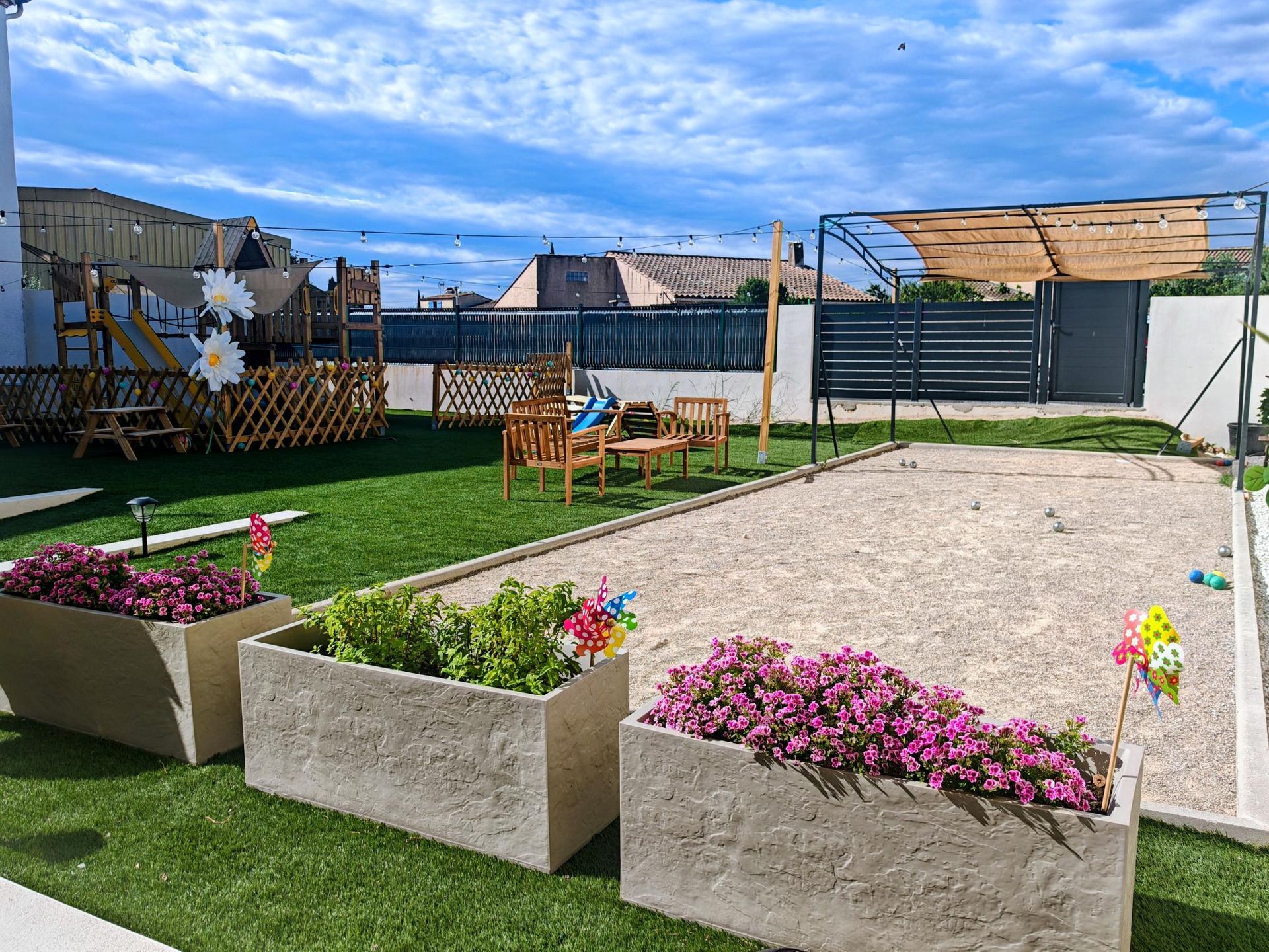 Patio de jardin avec parterres de fleurs surélevés, pelouse verte, table de pique-nique et coin salon en gravier sous un ciel bleu