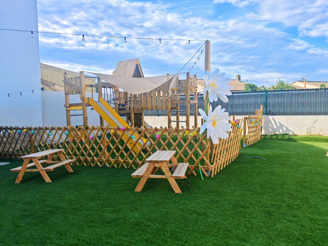 Aire de jeux avec clôture en bois, tables de pique-nique et grandes fleurs blanches sur une pelouse d'un vert éclatant sous un ciel bleu