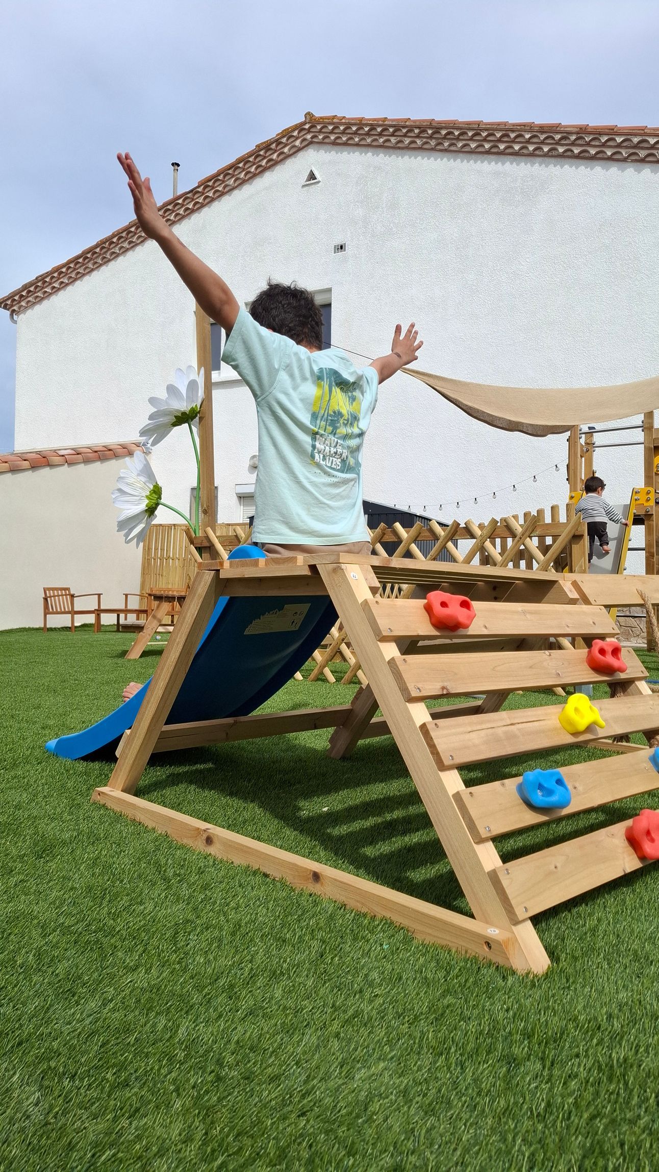 Une personne escalade une rampe de jeu en bois à l'extérieur, les bras levés, avec des prises colorées sur du gazon artificiel.