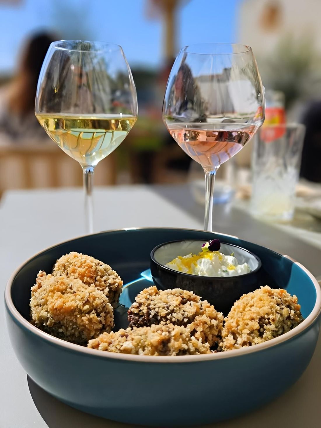 Des verres à vin derrière un bol de beignets panés avec une sauce, sur une table en terrasse.