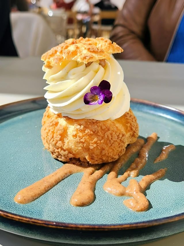 Pâtisserie garnie de crème fouettée et de fleurs violettes, présentée sur une assiette bleue et nappée de caramel.