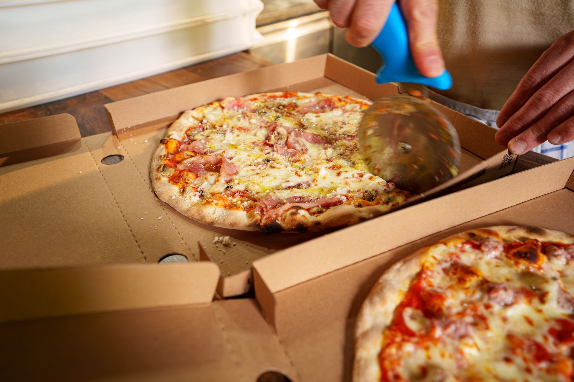 Deux pizzas dans des boîtes ouvertes, l'une d'elles coupée avec une roulette à pizza à manche bleu