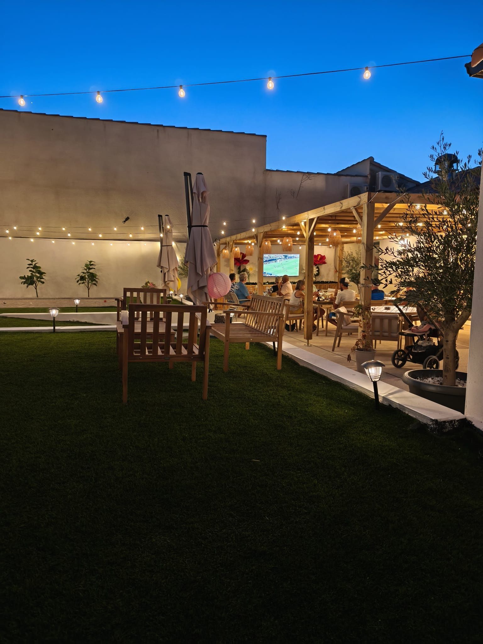 Des guirlandes lumineuses illuminent une agréable terrasse extérieure au crépuscule, avec des tables, des chaises et des plantes à côté d'un bâtiment.