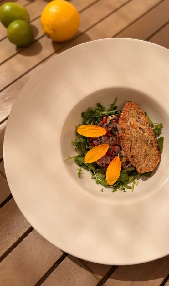 Plat gastronomique garni de verdure et d'orange, présenté sur une assiette blanche, sur une table en bois avec des citrons verts à proximité.