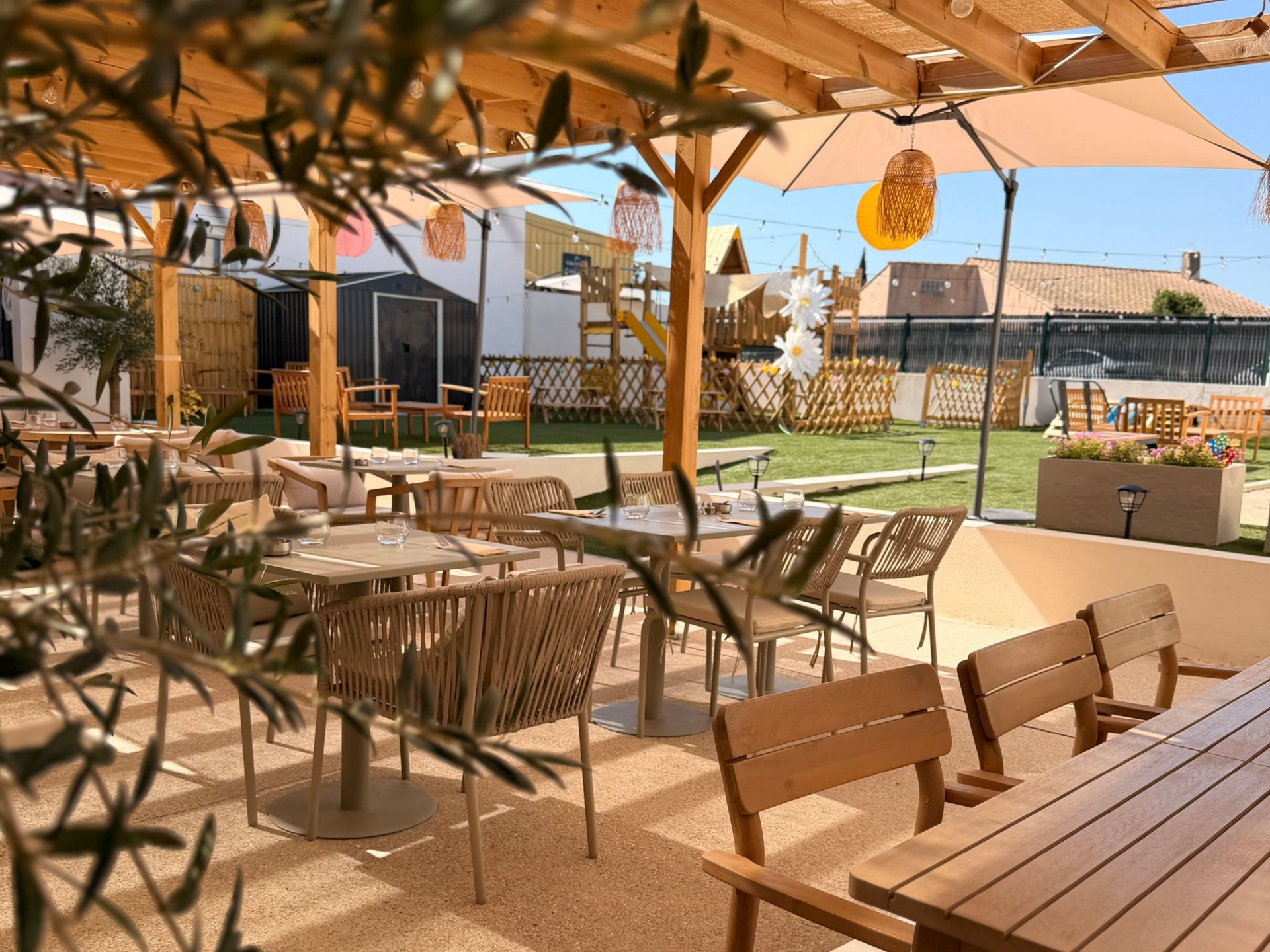 Terrasse de café en plein air avec tables et chaises en bois sous une pergola, donnant sur une cour gazonnée et des maisons