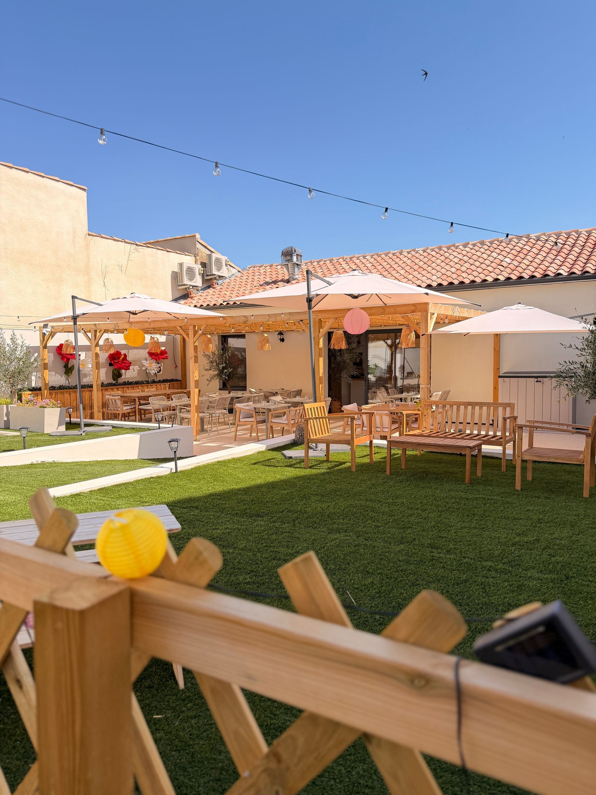 Cour intérieure ensoleillée avec tables en bois, parasols et bâtiments aux toits de tuiles rouges derrière un patio gazonné.