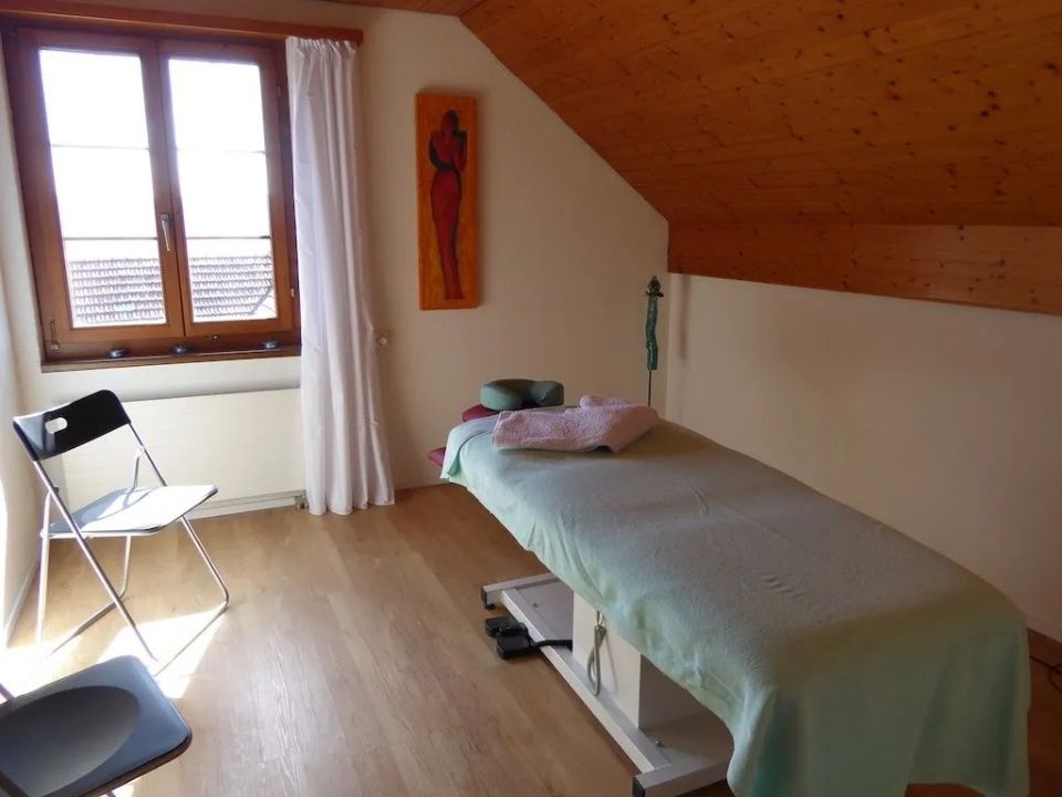 Massageraum mit Behandlungsliege, Holzdecke, Fenster mit weissen Vorhängen und einem Stuhl auf hellem Holzboden.