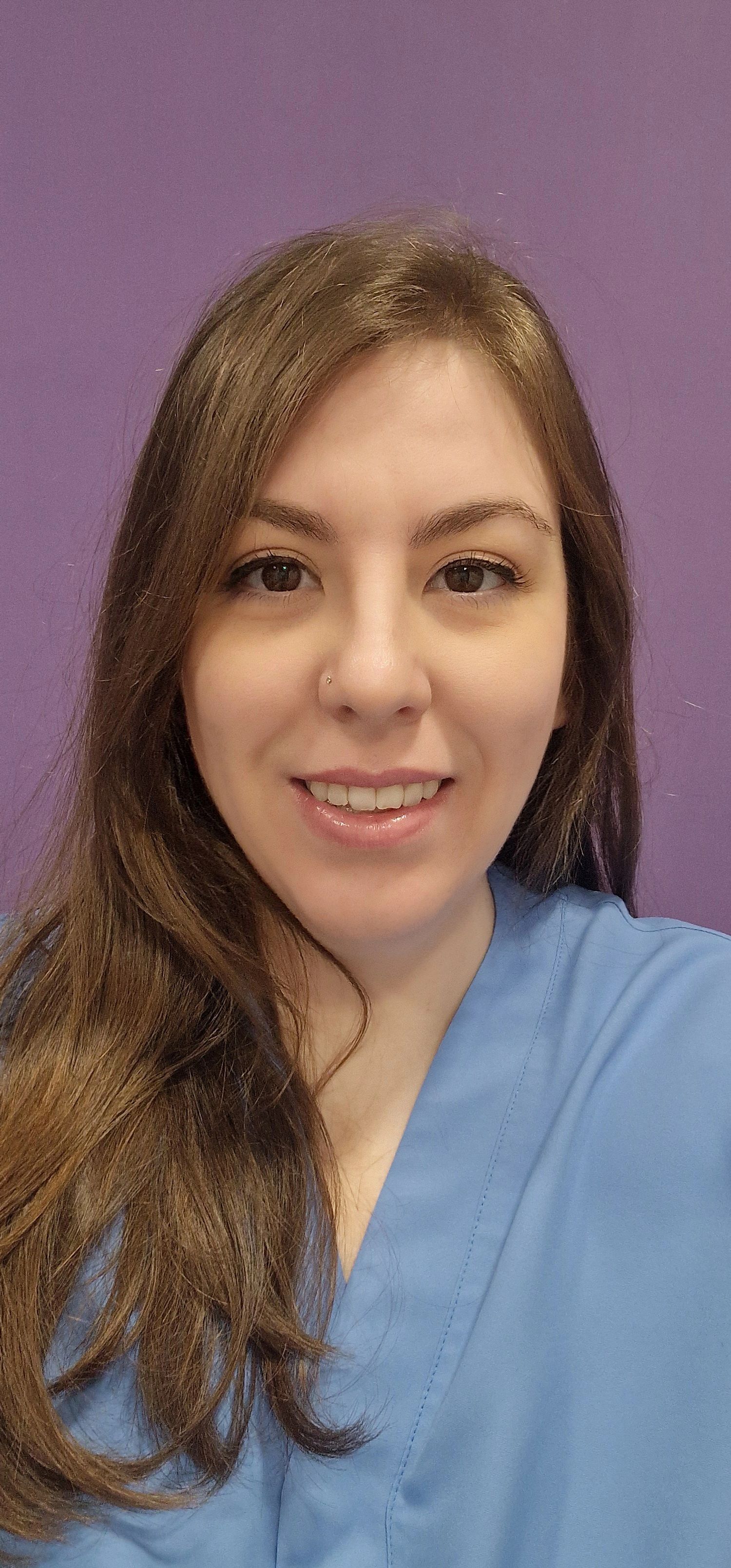 Una mujer que lleva gafas y una blusa azul sonríe frente a una pared violeta.