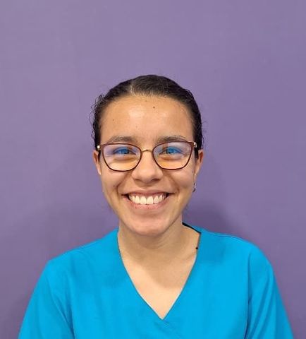 Una mujer que lleva gafas y una blusa azul sonríe frente a una pared violeta.