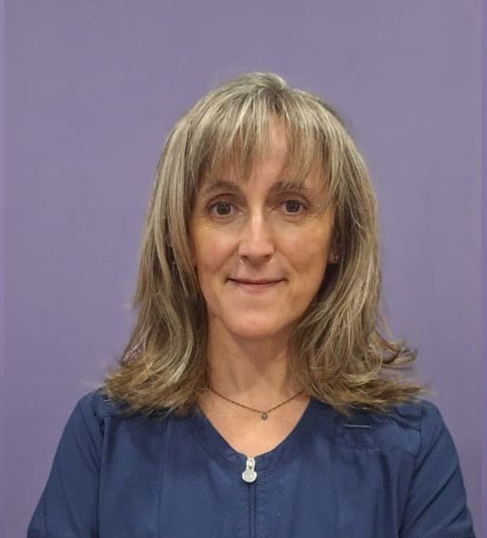 Una mujer que viste una blusa azul y un collar está parada frente a una pared violeta.
