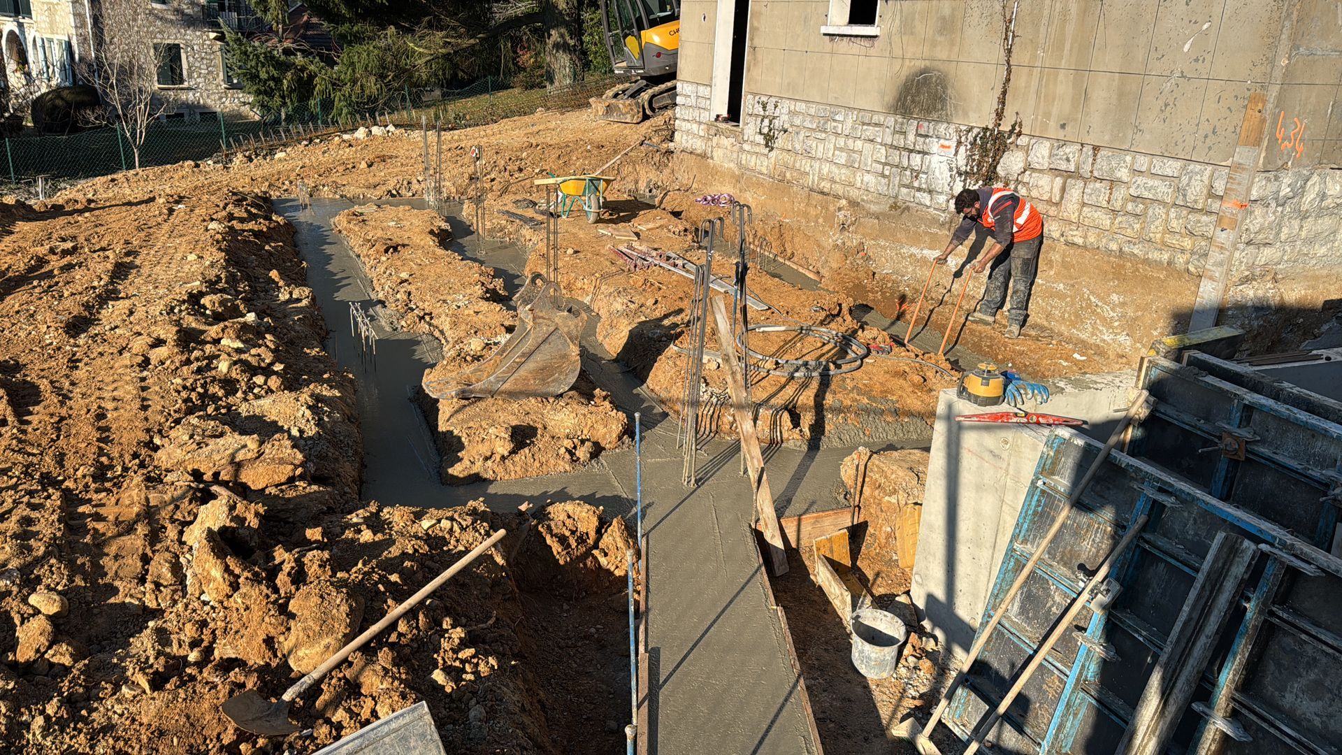 Chantier de construction avec tranchées creusées, mur en béton et deux ouvriers.