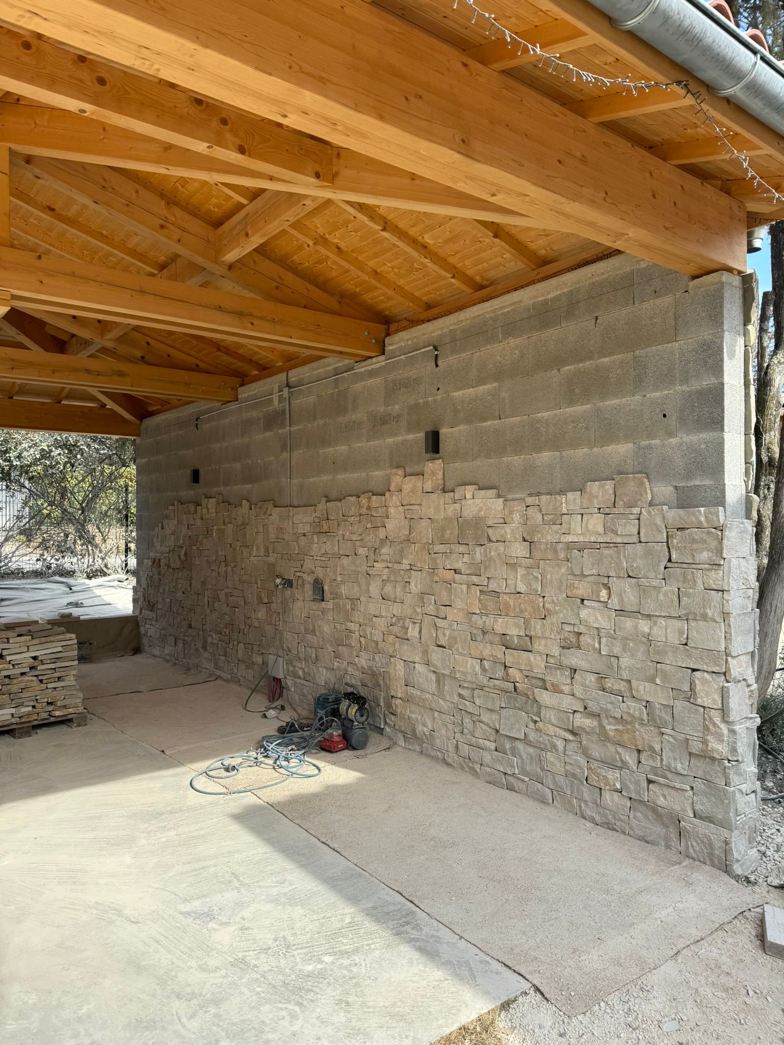 Un mur en pierre sous une charpente en bois, avec un sol en béton.