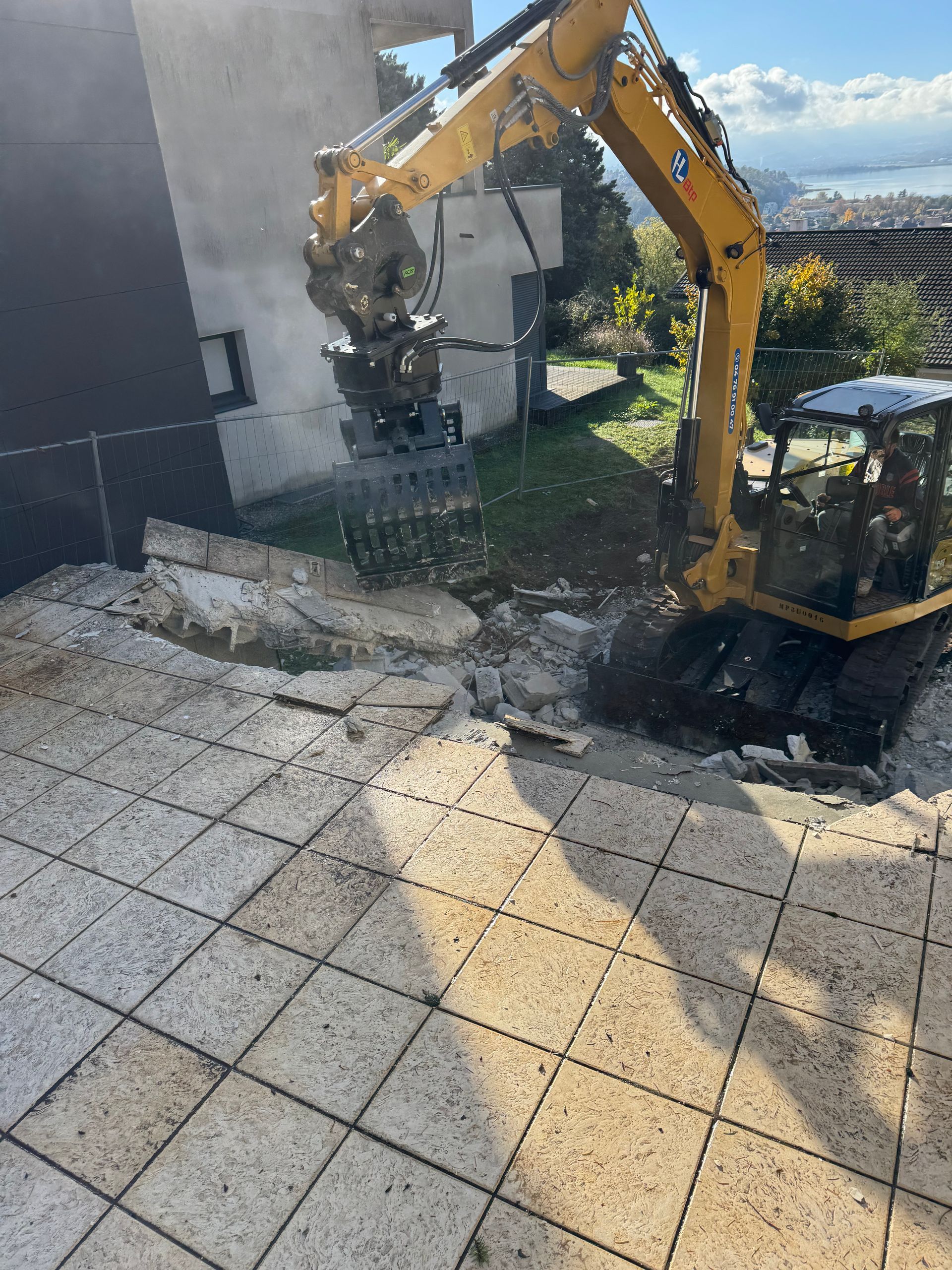 Une pelleteuse jaune démolit un mur en béton à côté d'une terrasse carrelée.