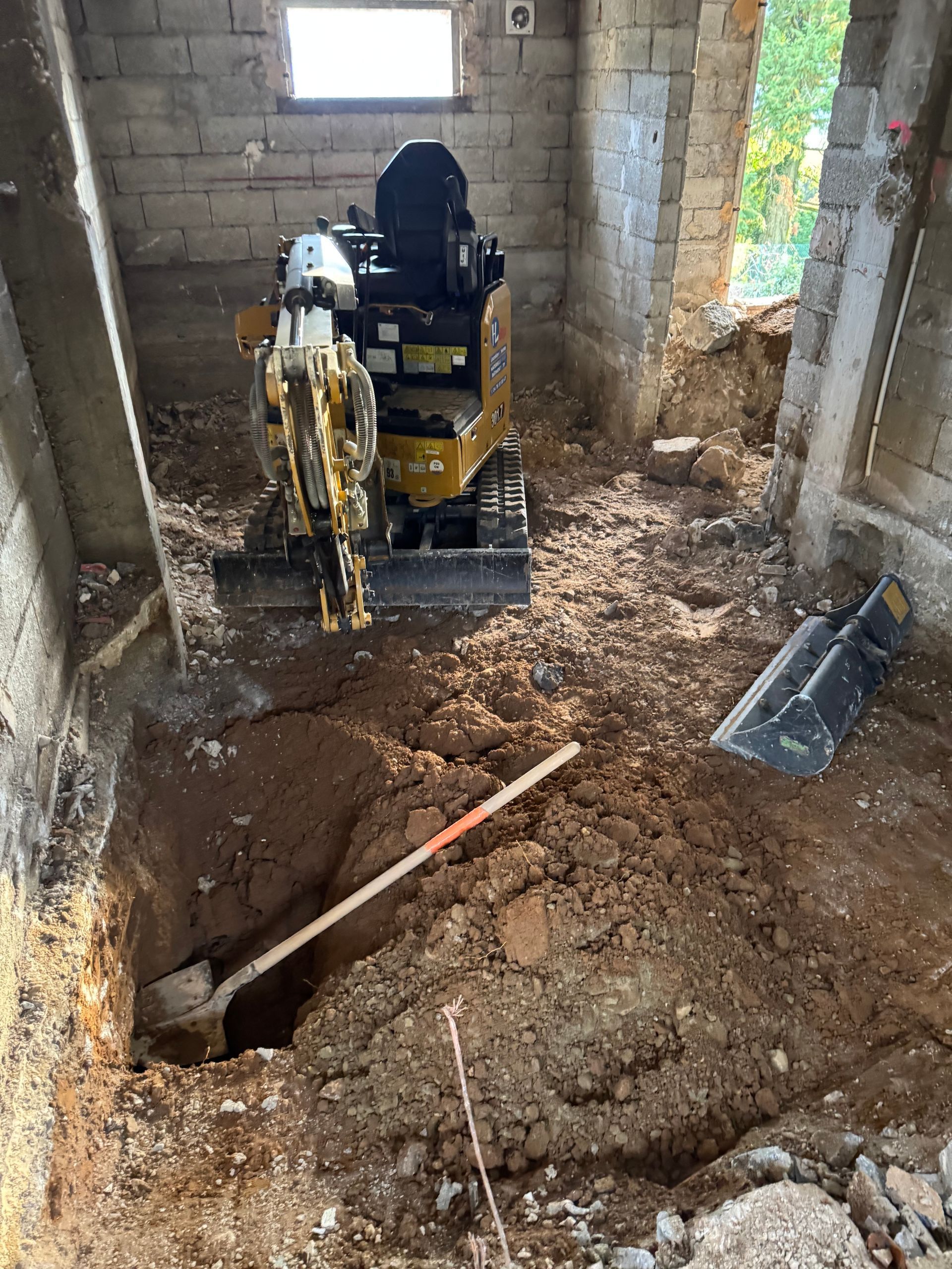 Chantier avec une excavatrice creusant une tranchée dans un sol en terre battue sous une fenêtre.