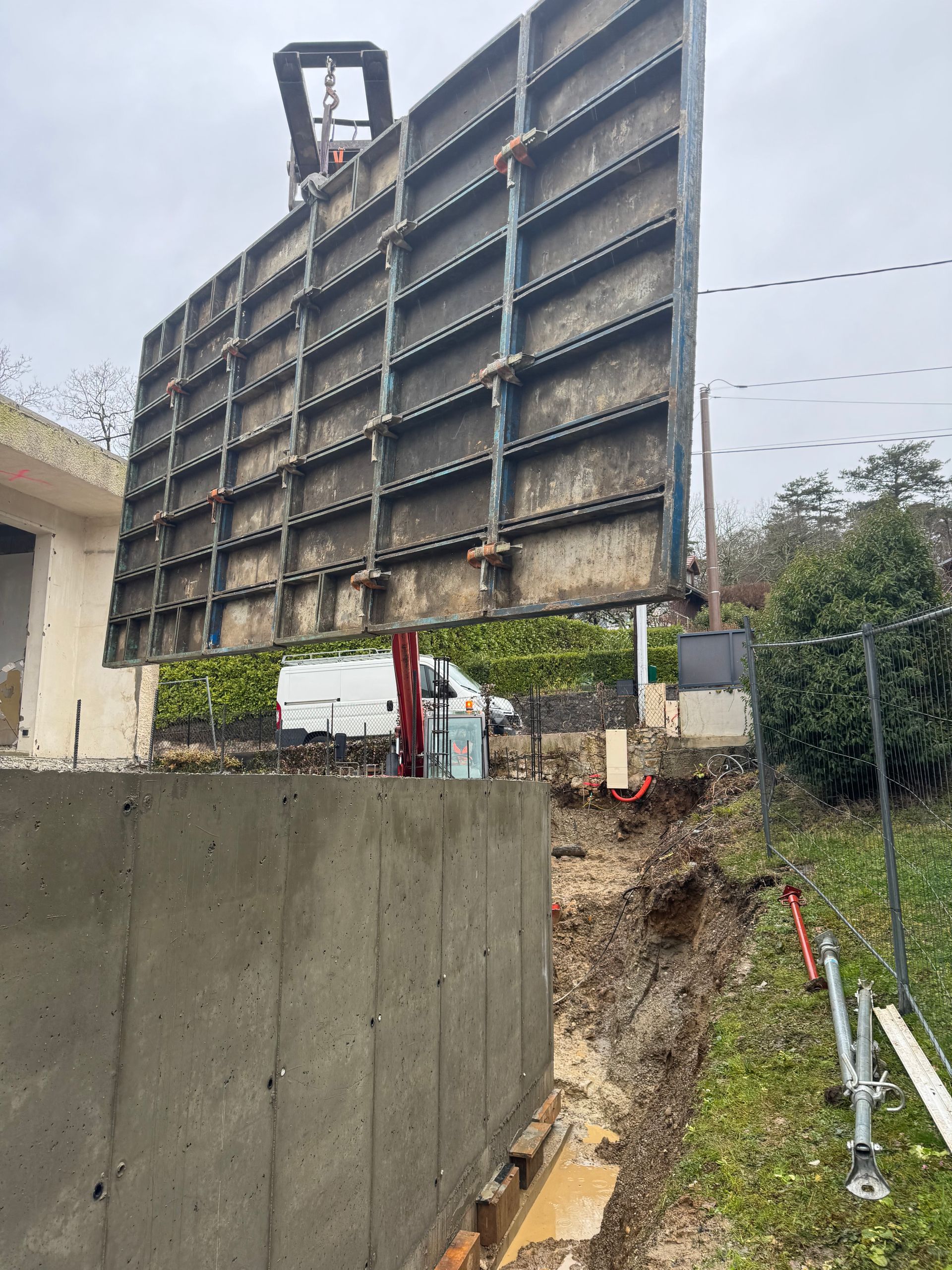 Un grand panneau de coffrage en acier pour béton suspendu par une grue au-dessus d'un mur en béton.
