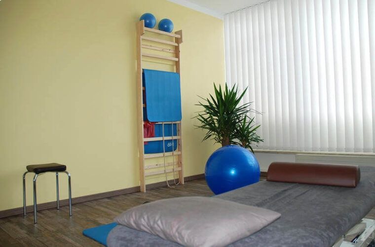 Ruhiger Raum mit Massageliege, Kissen, blauem Gymnastikball, Pflanze und an der Wand befestigter Leiter.