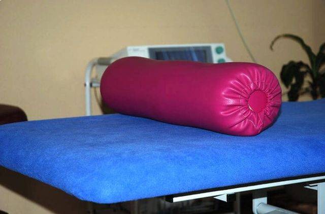 Ein rosa zylinderförmiges Kissen auf einem blauen gepolsterten Tisch in einem Klinik- oder Therapieraum.
