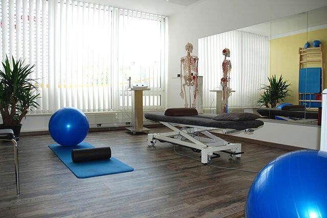 Physiotherapieraum mit Behandlungsliege, Gymnastikbällen, Matte, Skelettmodellen und hellen Fenstern