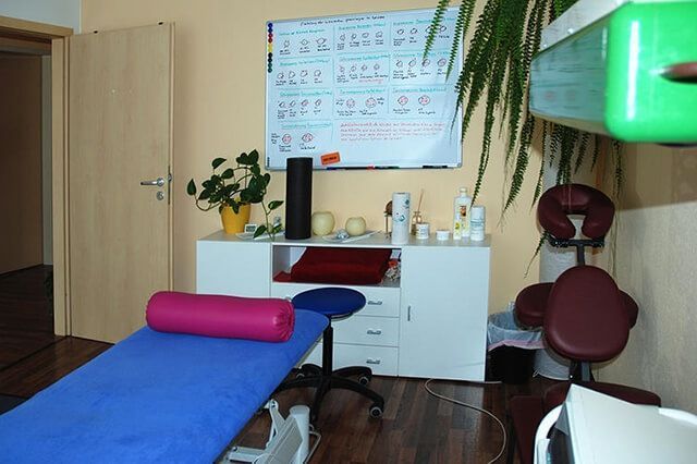 Massageraum mit blauer Behandlungsliege, rotem Hocker, weißem Schrank und Wandtafel