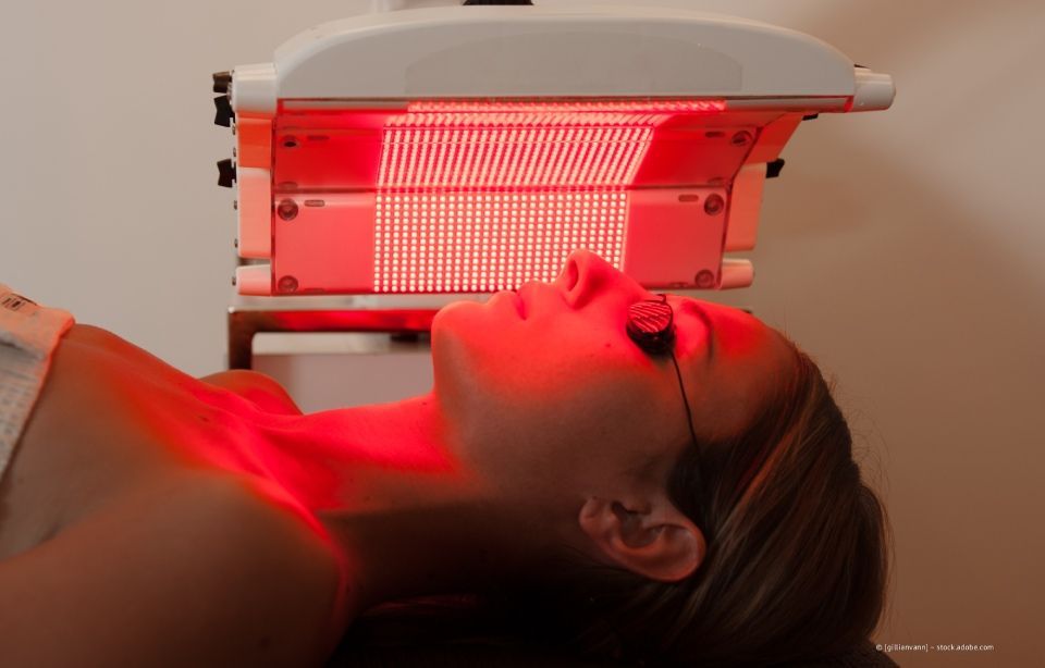 Eine Person liegt unter einer Rotlichttherapie-Lampe; Gesicht und Hals leuchten rot.