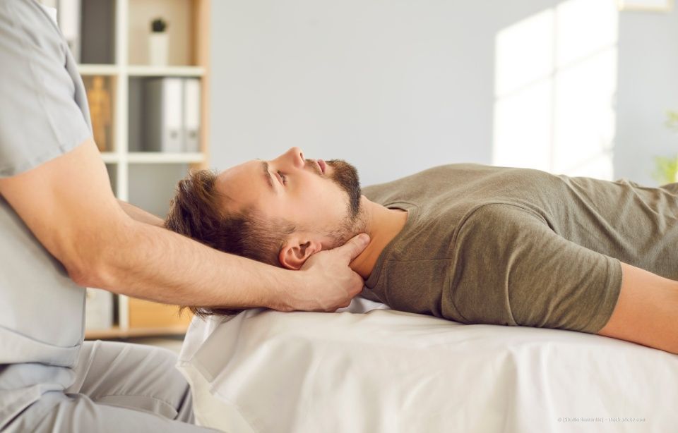 Eine Massagetherapeutin massiert den Nacken eines Klienten auf einer Behandlungsliege in einem hellen Raum.