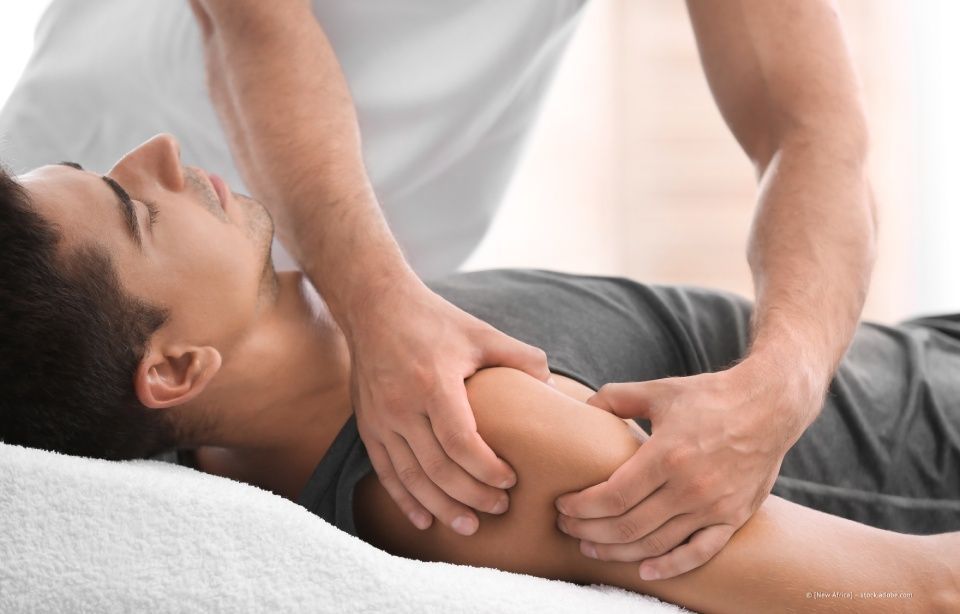 Ein Physiotherapeut massiert die Schulter eines Patienten, der auf einer weißen Behandlungsliege liegt.