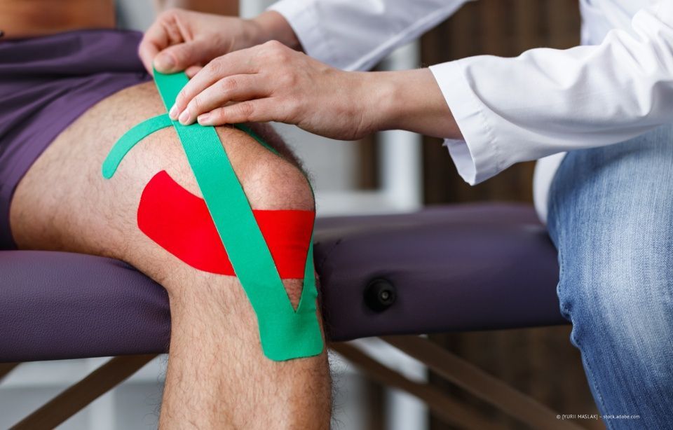 Knie-Taping während der Physiotherapie, wobei grünes Kinesiologie-Tape auf ein gebeugtes Bein aufgebracht wird