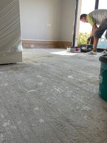 Persona pintando una habitación con piso de concreto, contenedores de pintura visibles.