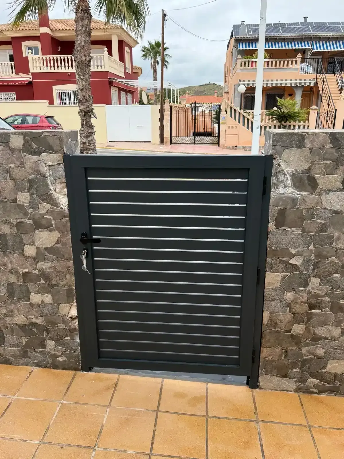 Puerta de metal negro en un muro de piedra, que da a un patio embaldosado y a casas residenciales detrás.