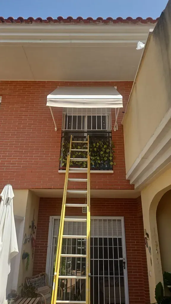 Una escalera amarilla apoyada contra una ventana del segundo piso de una casa de ladrillo rojo con toldo blanco.