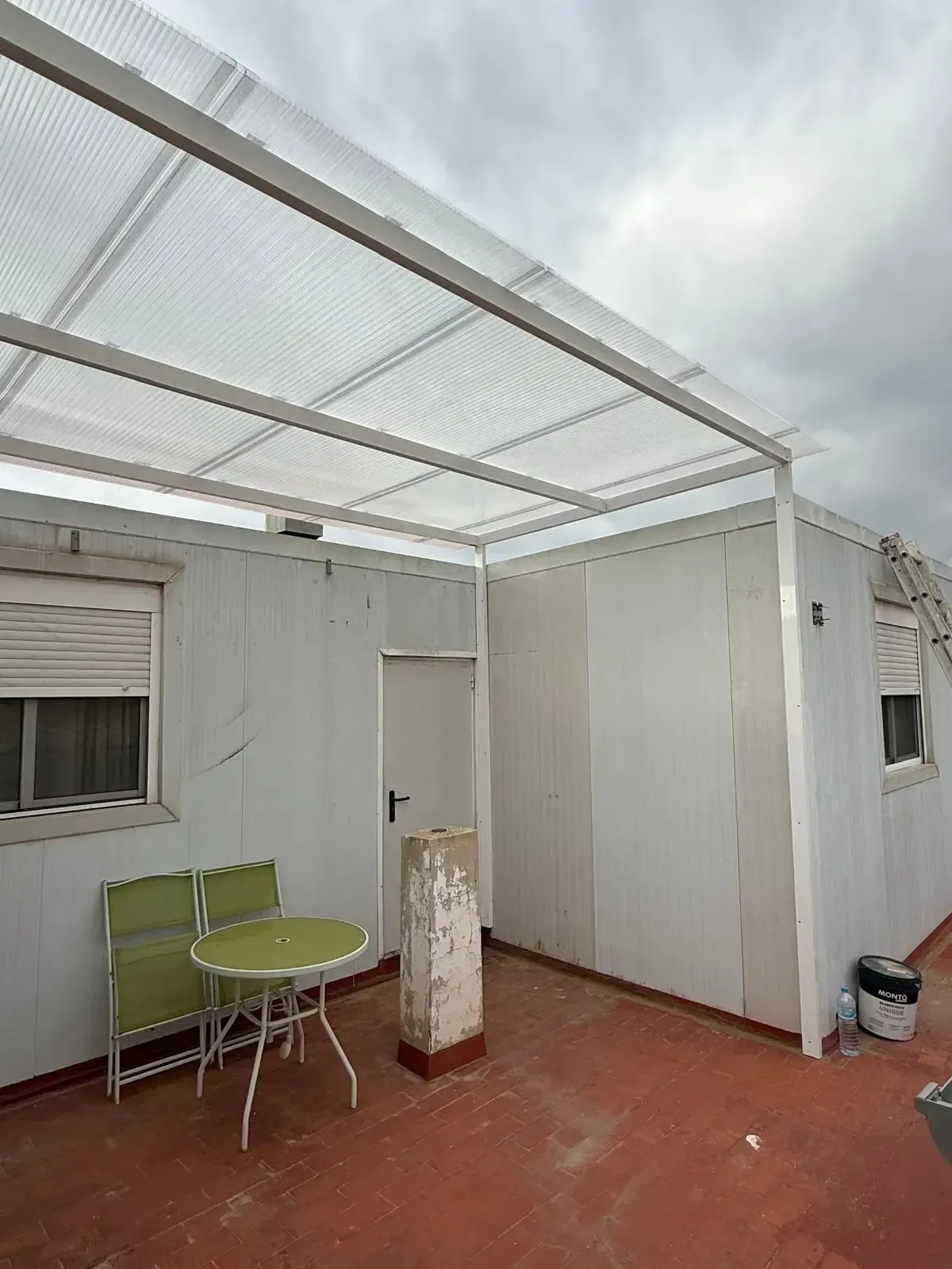 Pequeño patio blanco con suelo rojo, sillas verdes, mesa de cristal y techo con mosquitera.