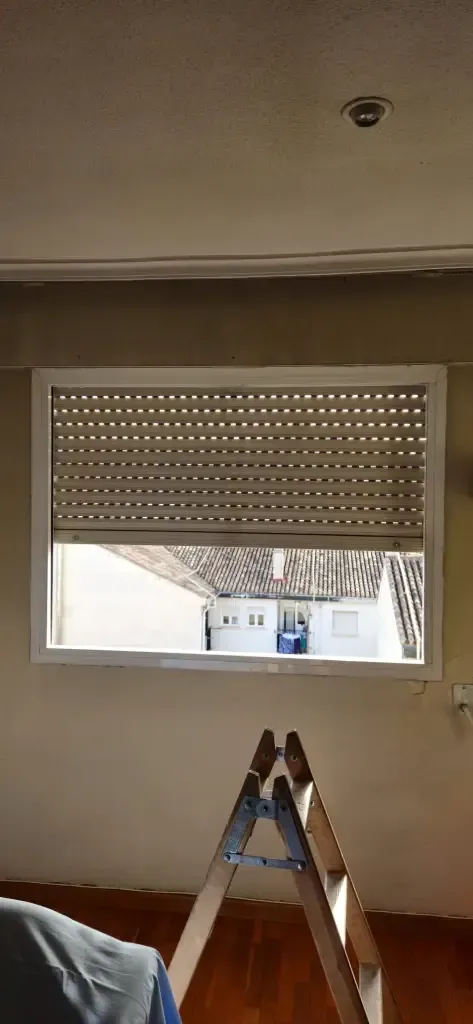 Habitación con una ventana abierta y una escalera en primer plano, que deja ver una persiana enrollable parcialmente subida.
