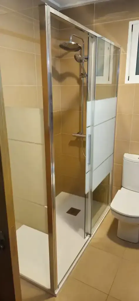 Cabina de ducha con mampara de cristal y azulejos beige, con un inodoro al lado, en un baño.