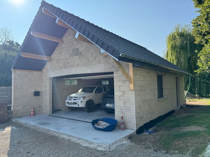 Garage avec deux voitures garées à l'intérieur, briques de couleur claire et toit en angle avec poutres en bois.
